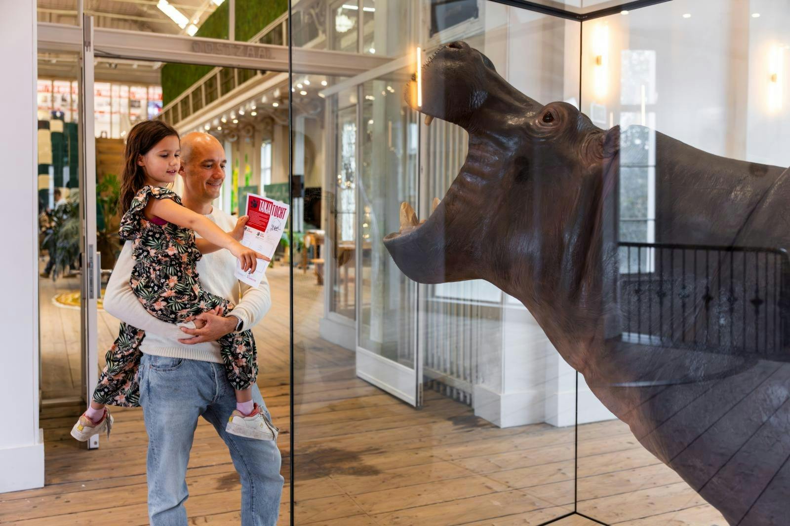 Un homme tient un enfant devant un présentoir en verre contenant une sculpture d'hippopotame. L'enfant tient une brochure et pointe du doigt le présentoir.