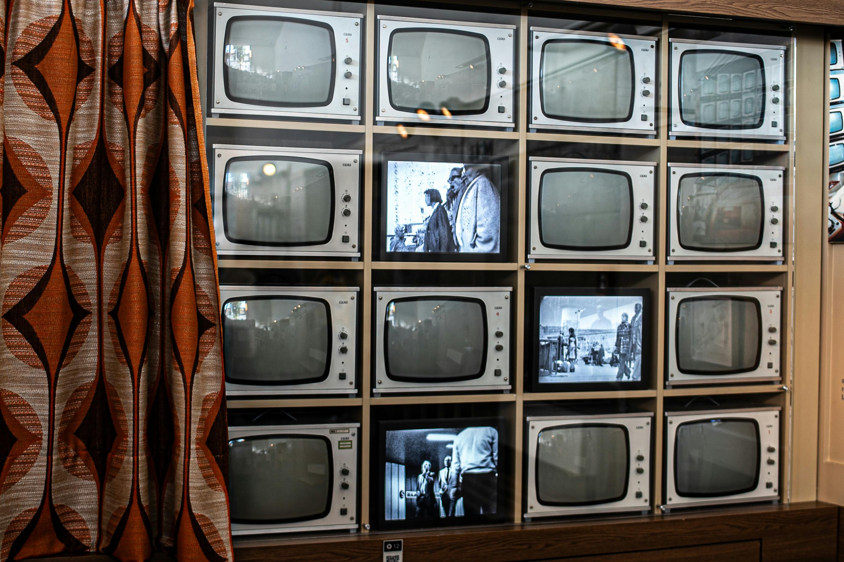 Uma parede com televisores antigos, alguns com cenas a preto e branco, emoldurada por uma cortina com padrões à esquerda.