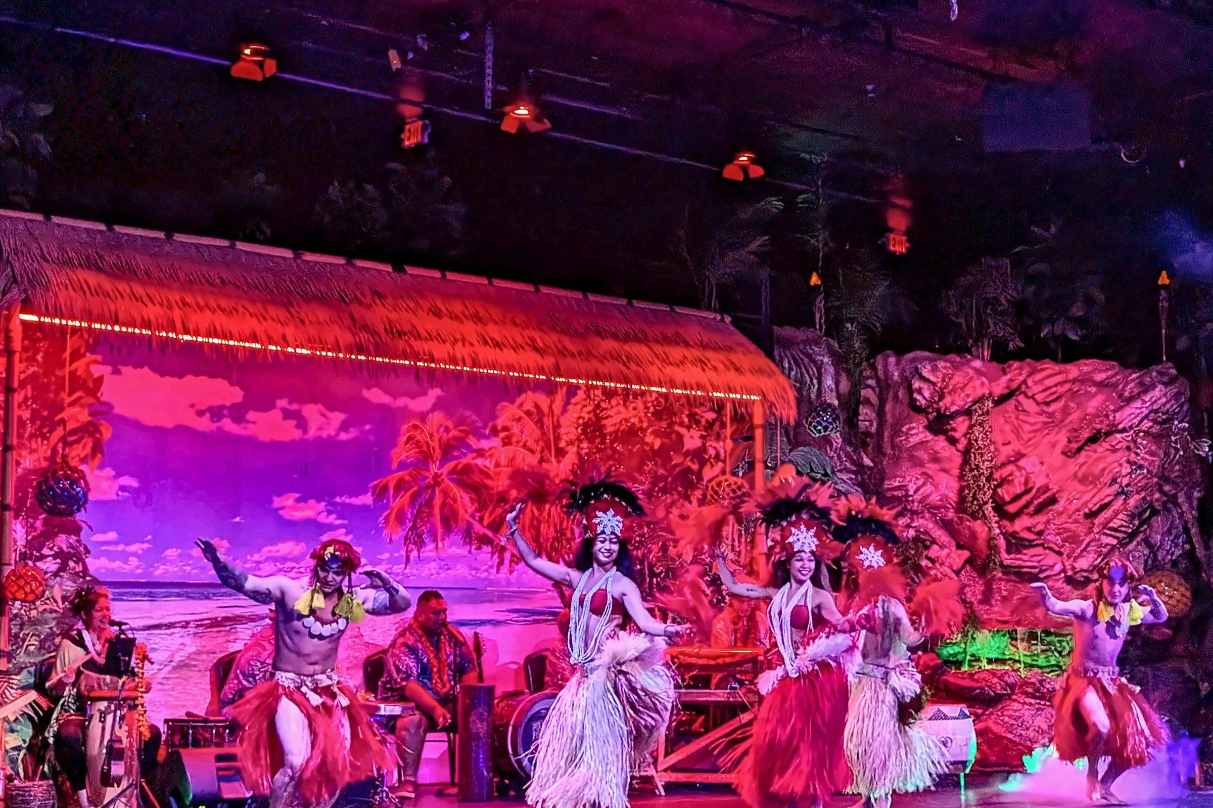 A Polynesian dance performance with dancers in traditional attire on a stage with tropical-themed decorations.