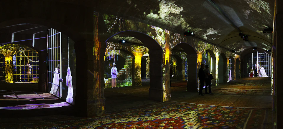 An illuminated stone archway with projected art, featuring a figure in a purple robe and a person walking on a patterned floor.
