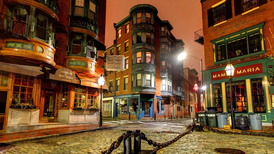 Illuminated street corner with historic brick buildings, storefronts, and cobblestone road at twilight. Chains and lamppost in the foreground.