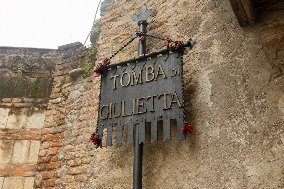 A metal sign with the text 'Tomba di Giulietta' hangs on a pole against a textured wall and brick background.