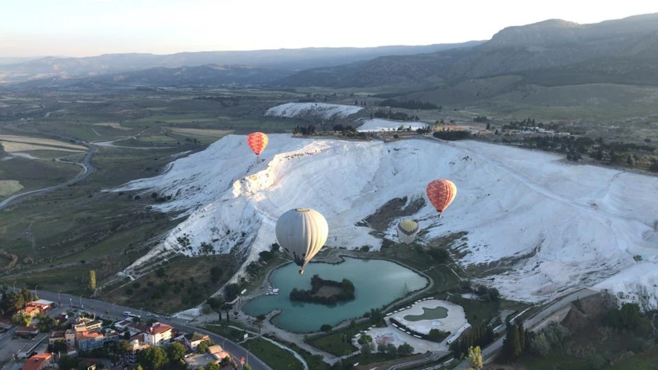 Three hot air balloons float over white limestone terraces and turquoise pools with mountains in the distance.