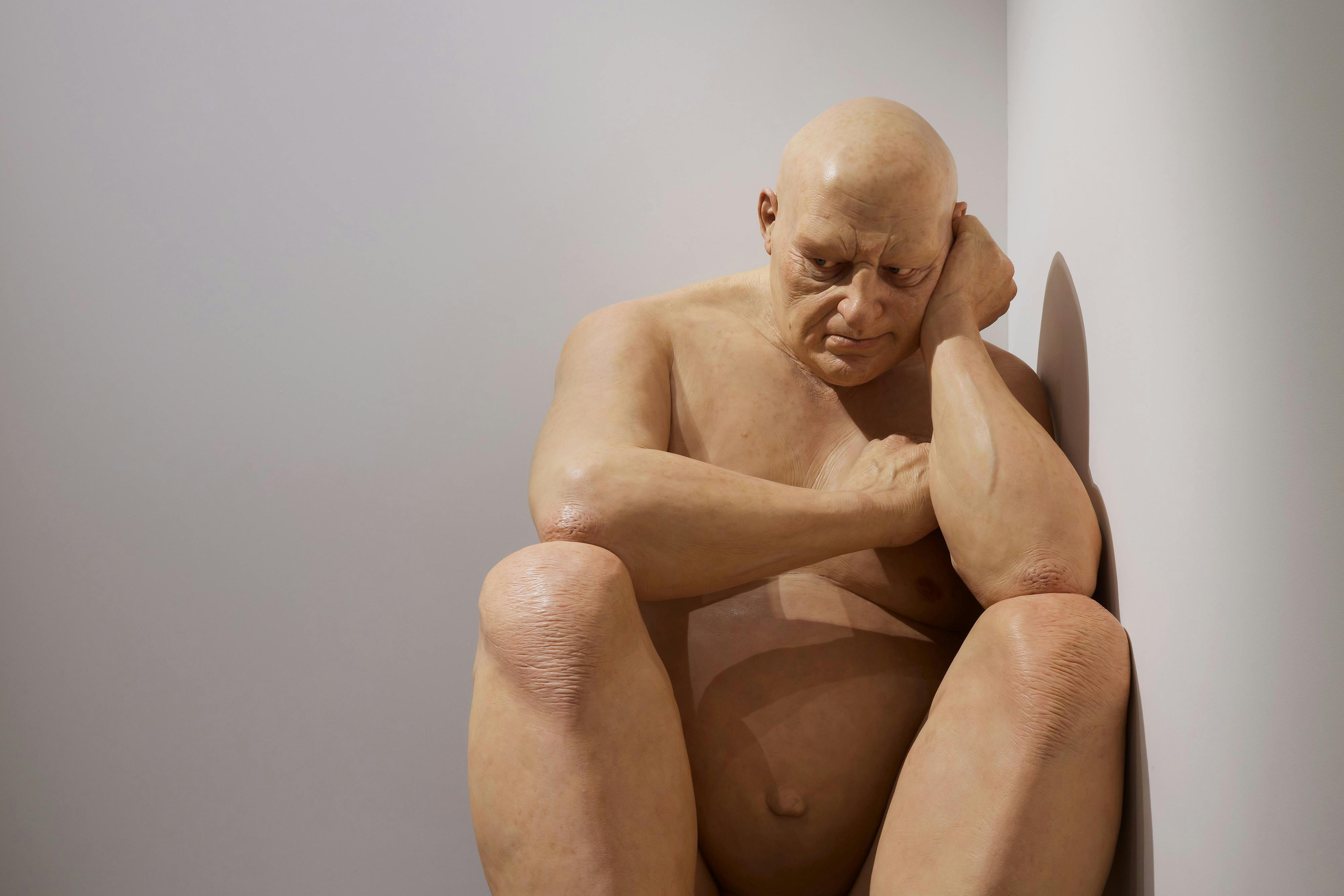A hyper-realistic sculpture of a seated, bald, and unclothed man, positioned in a corner with a contemplative expression.