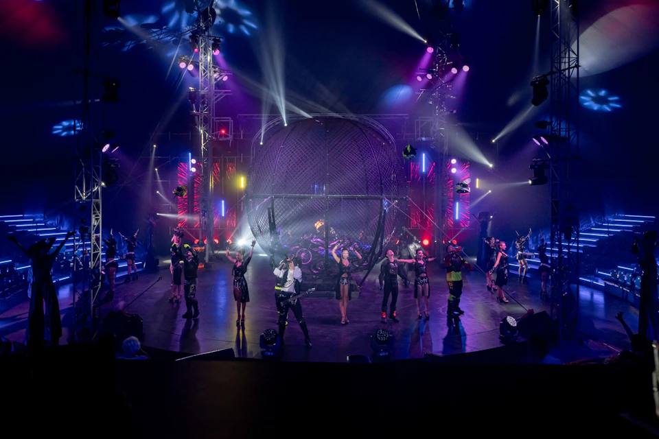 Performers on a brightly lit stage with a spherical cage centerpiece, colorful lights, and an audience in the background.