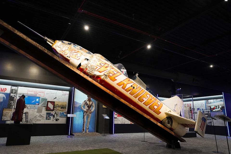 A rocket-like vehicle labeled 'Evel Knievel' displayed on a slanted platform in a museum setting with informational boards nearby.
