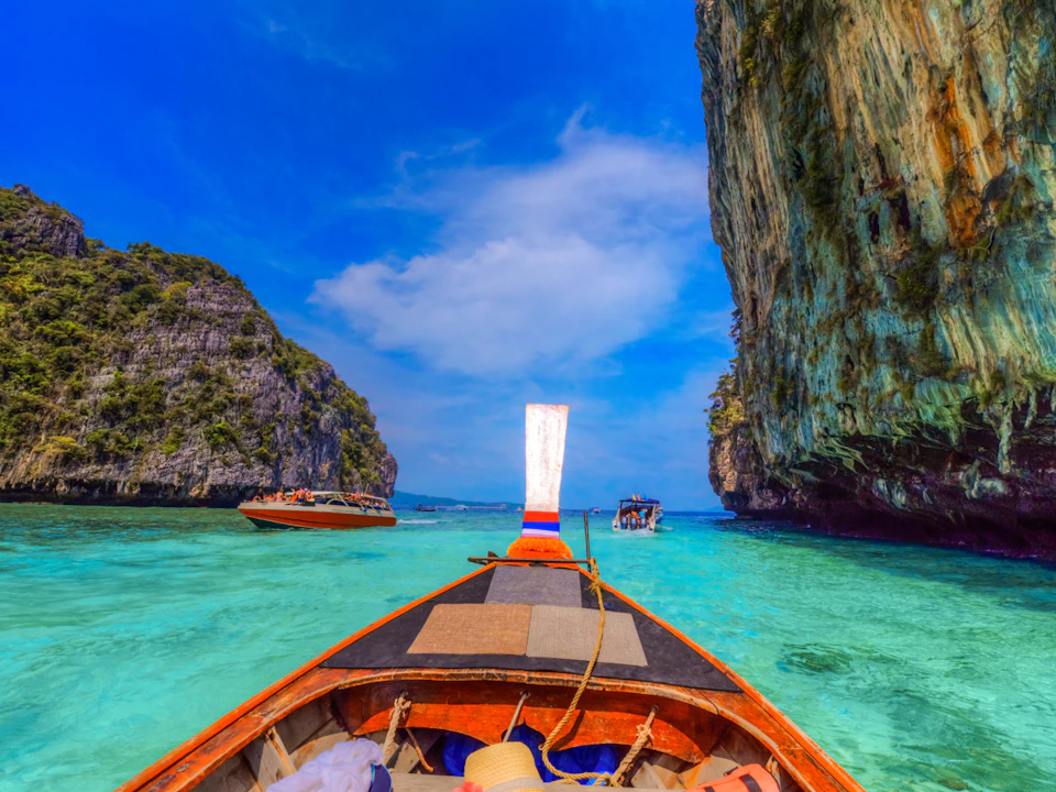 View from a wooden boat of clear turquoise water surrounded by tall cliffs and other boats beneath a bright blue sky.
