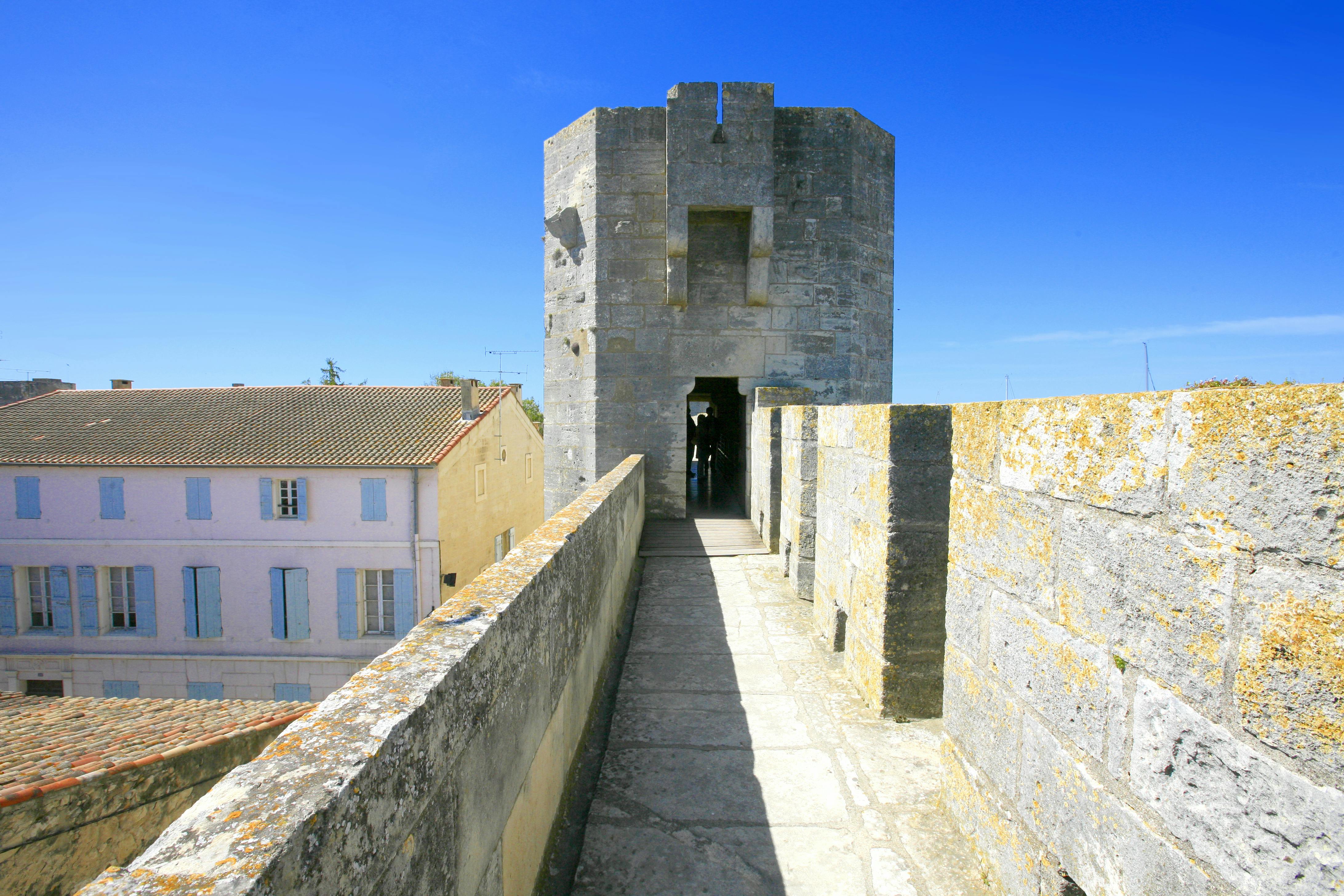 Stone walkway leads to a tower entrance with clear blue sky. Adjacent are rooftops of pastel-colored buildings.
