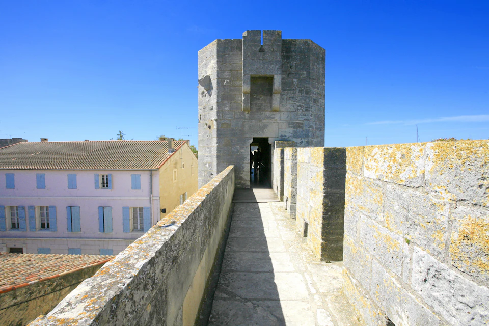 Stone walkway leads to a tower entrance with clear blue sky. Adjacent are rooftops of pastel-colored buildings.