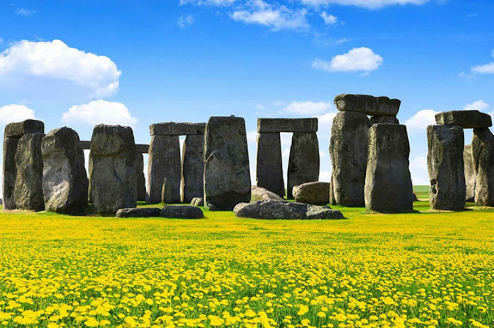 Stonehenge monument with large standing stones in the background, set in a field of blooming yellow flowers under a partly cloudy sky.
