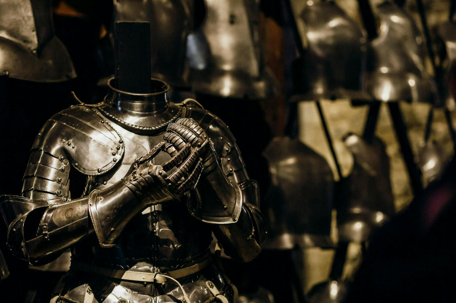 A suit of medieval armor with clasped gauntlets on display, surrounded by other armors in the background.