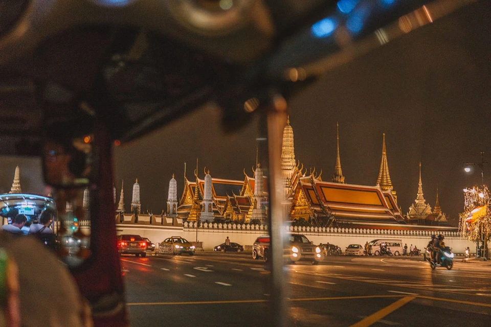 Nighttime view of a brightly lit, ornate temple complex with spires. Cars pass by on the adjoining road.