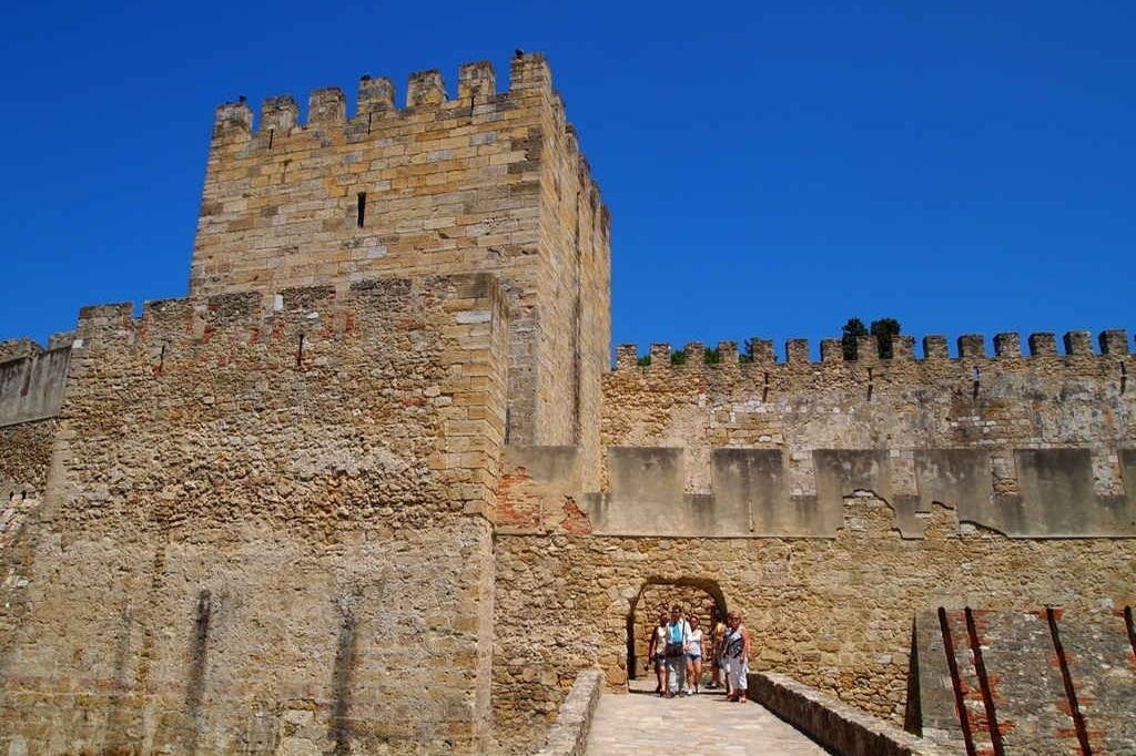People walking through a stone archway in a historic castle with battlements, under a clear blue sky.