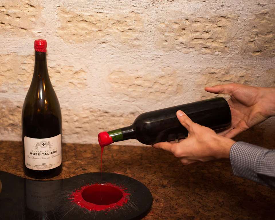 A person is coating a wine bottle neck with red wax near a table holding another wine bottle, against a stone wall.