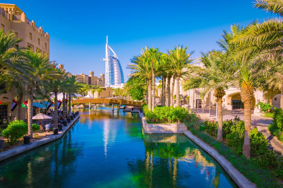 Modern skyscraper in the background, palm trees lining a blue canal with a bridge, and buildings in the foreground under a clear sky.