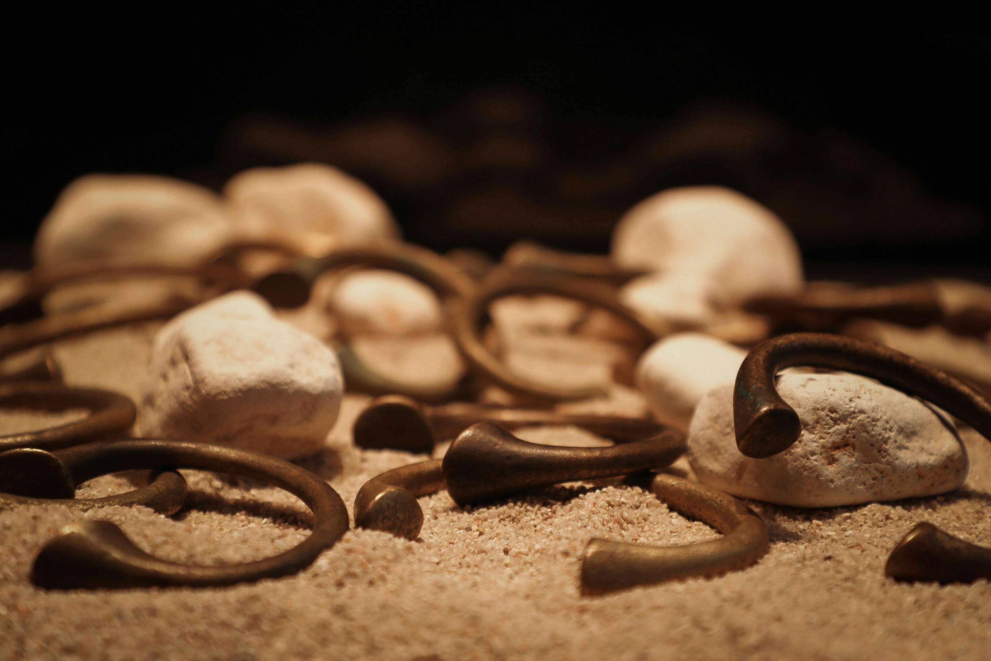 Δαχτυλίδια και κυλινδρικά αντικείμενα ακουμπούν σε μια αμμώδη επιφάνεια, που περιβάλλεται από λευκές πέτρες σε σκούρο φόντο.