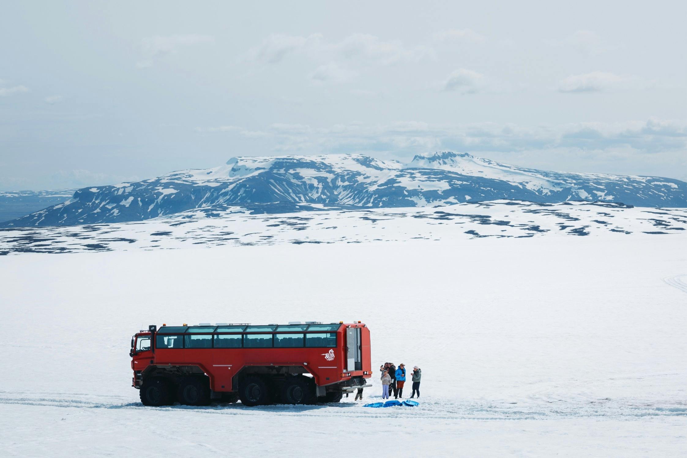Un veicolo rosso da ghiacciaio e diverse persone si trovano in un ampio paesaggio innevato con montagne innevate sullo sfondo.