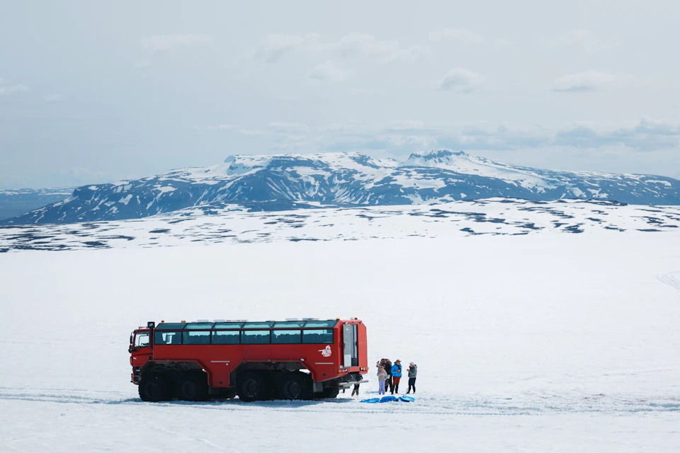 A red glacier vehicle and several people stand on an expansive snowy landscape with snow-covered mountains in the background.
