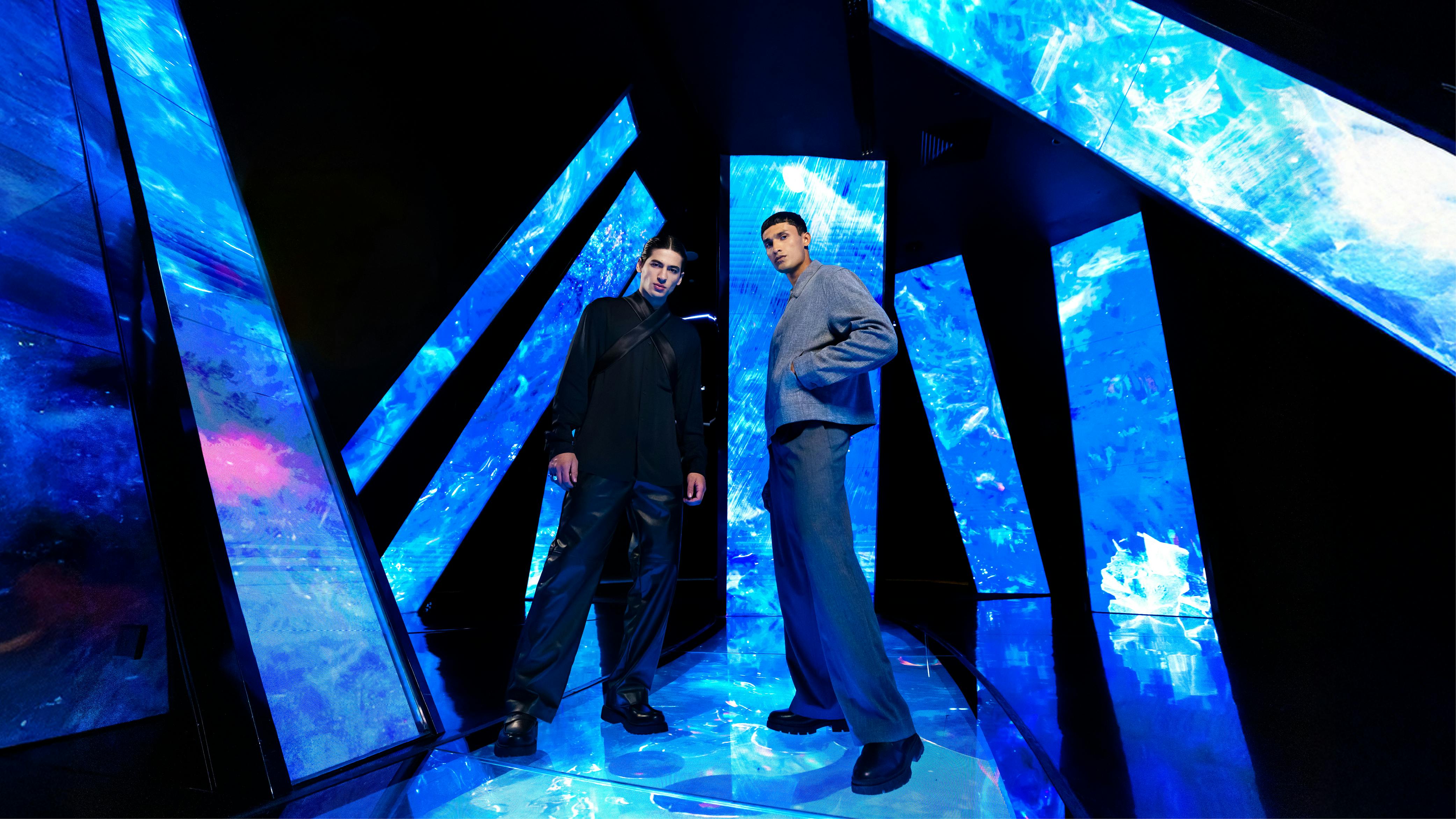 Δύο άνδρες ποζάρουν σε ένα φουτουριστικό σκηνικό με μεγάλα γεωμετρικά πάνελ μπλε φωτός. Είναι ντυμένοι με σκούρα, μοντέρνα ρούχα.