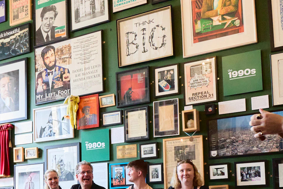A group of four people sit on a bench in a room with many framed photos, posters, and artwork on the wall behind them.