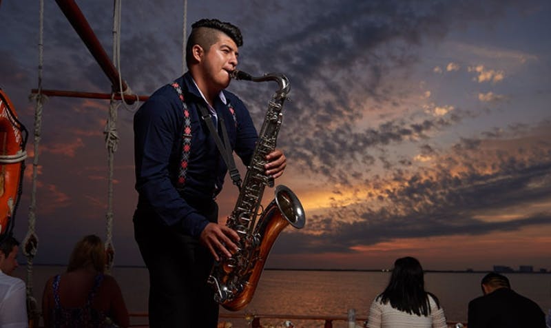 Un uomo che suona un sassofono su una barca al tramonto, con un'altra persona seduta sullo sfondo. Il cielo è pieno di nuvole drammatiche.