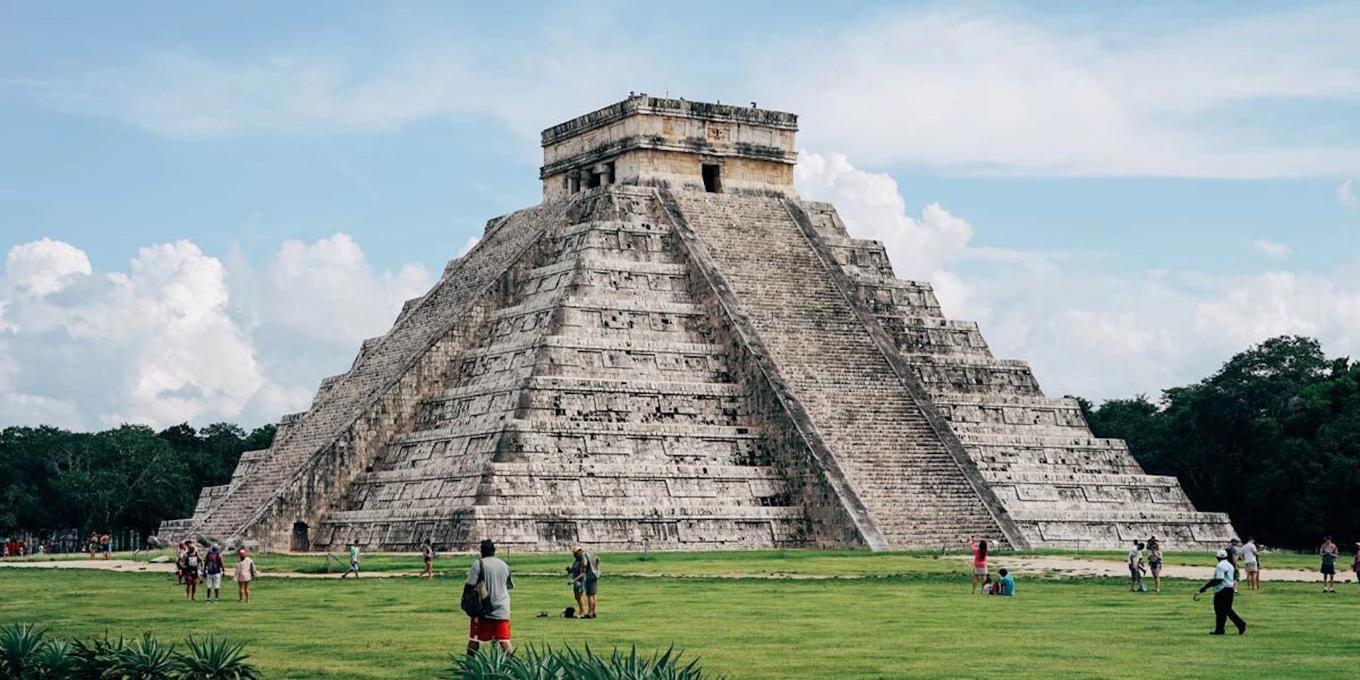 A large stone pyramid with steep steps in a grassy area, with several people walking and observing the structure.