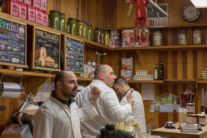 Three men in white coats work behind a deli counter with jars and chalkboard menus on the wall.