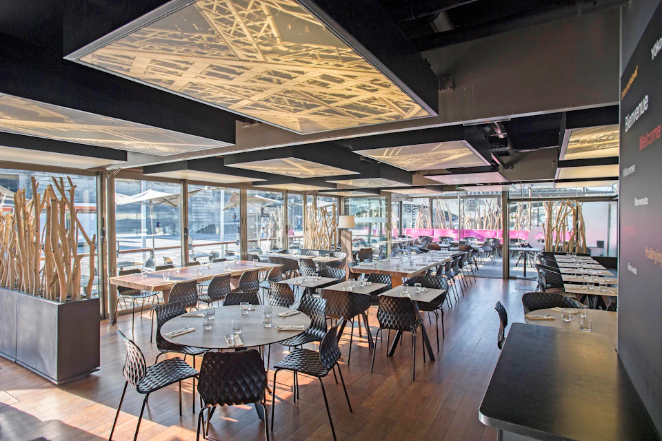 A modern restaurant with empty black chairs and round tables, illuminated ceiling panels, and large windows offering outside views.