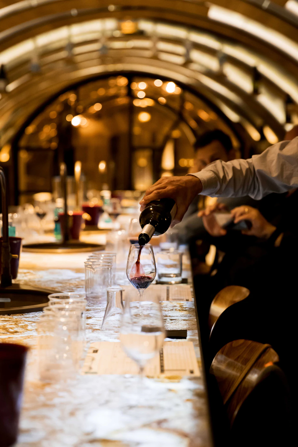 A person pours red wine into a glass on a long, well-lit table with rows of glasses in a cozy, arched interior setting.