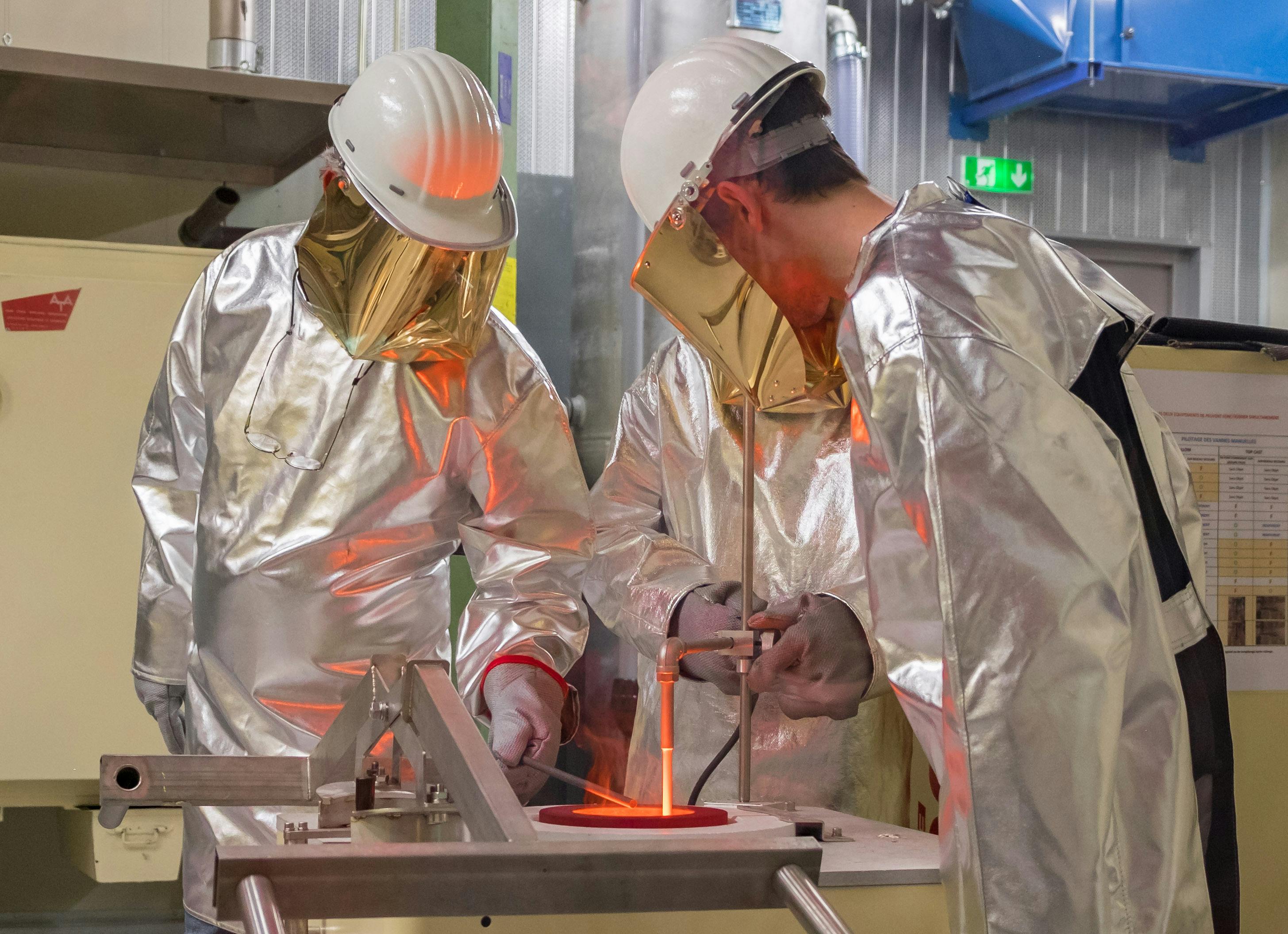 Δύο εργάτες με ασημένιες προστατευτικές στολές και κράνη διαχειρίζονται λιωμένο μέταλλο σε βιομηχανικό χυτήριο.