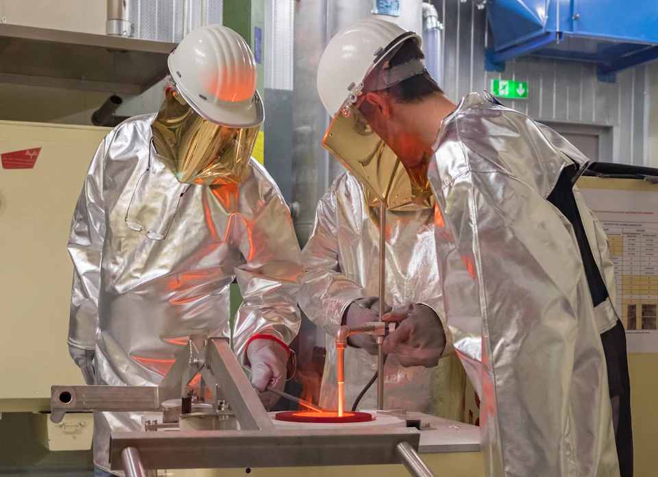 Two workers in silver protective suits and helmets manage molten metal in an industrial foundry setting.