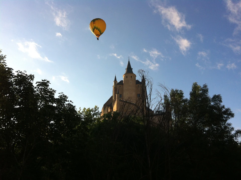 A hot air balloon floats in the sky near a castle with spires, surrounded by trees, under a partly cloudy sky.