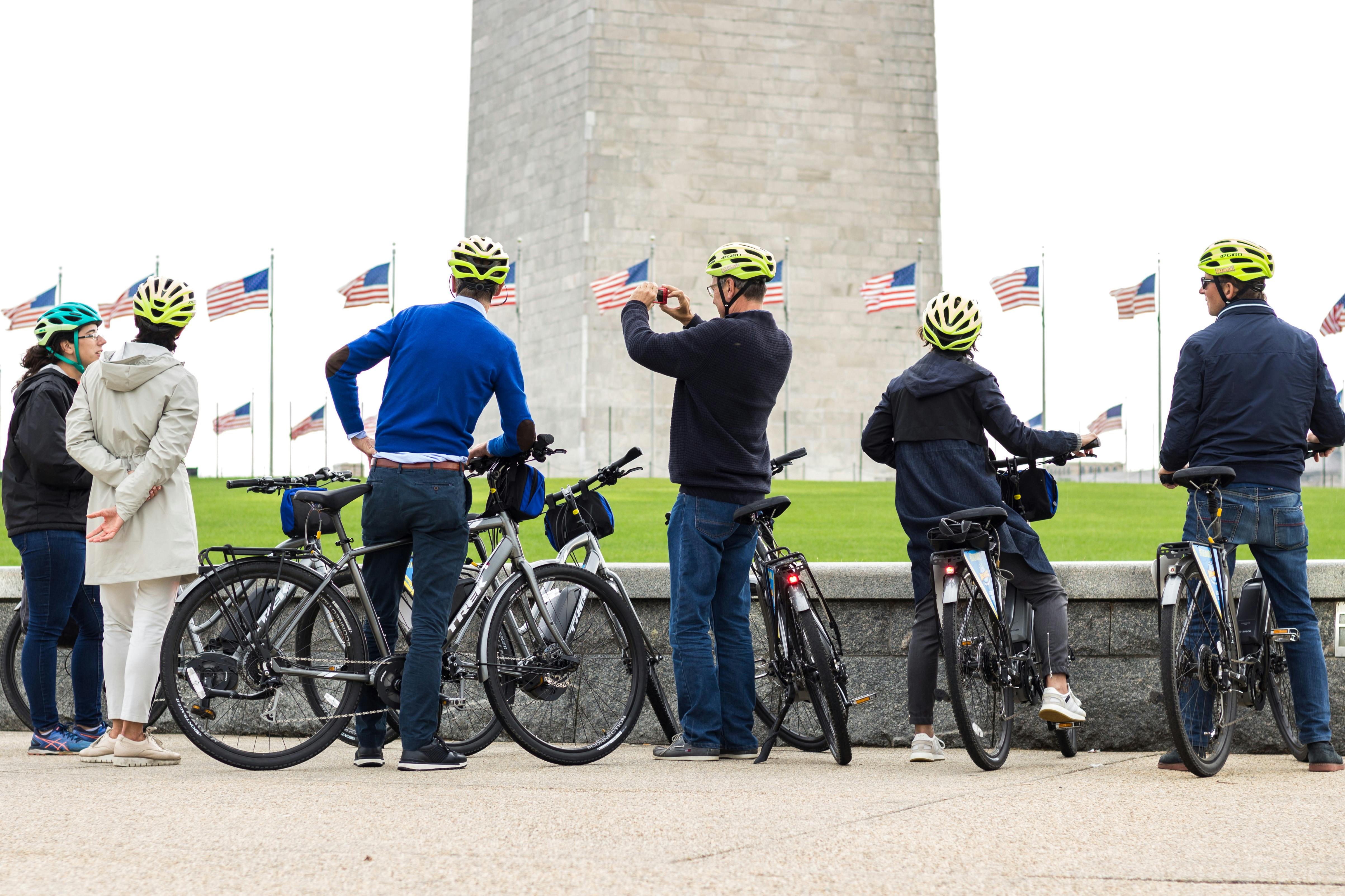 Un gruppo di ciclisti con il casco sta in piedi vicino alle loro biciclette, guardando le bandiere davanti a un grande monumento di pietra.