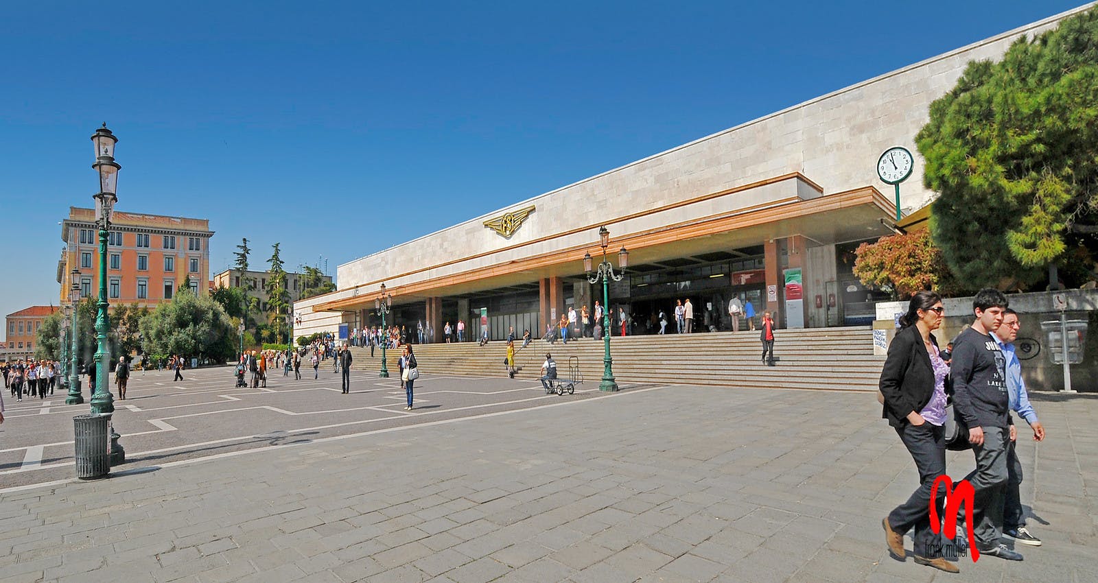 Menschen gehen auf einem großen Platz vor einem großen Gebäude mit Stufen, einer Uhr und einem Schild mit einem geflügelten Emblem.