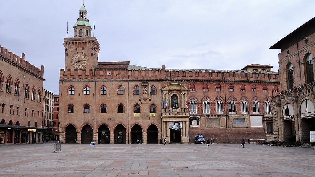 A large plaza with medieval buildings, including a clock tower and arcades, with a few people walking around.