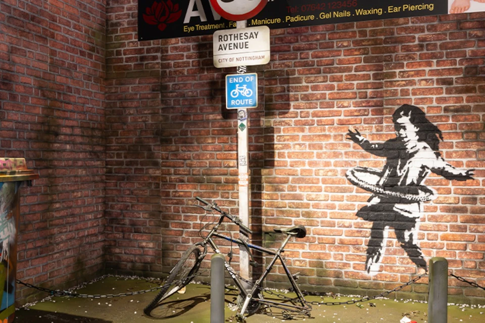 A bicycle rests against a pole with signs in front of a brick wall with graffiti of a hula-hooping figure.