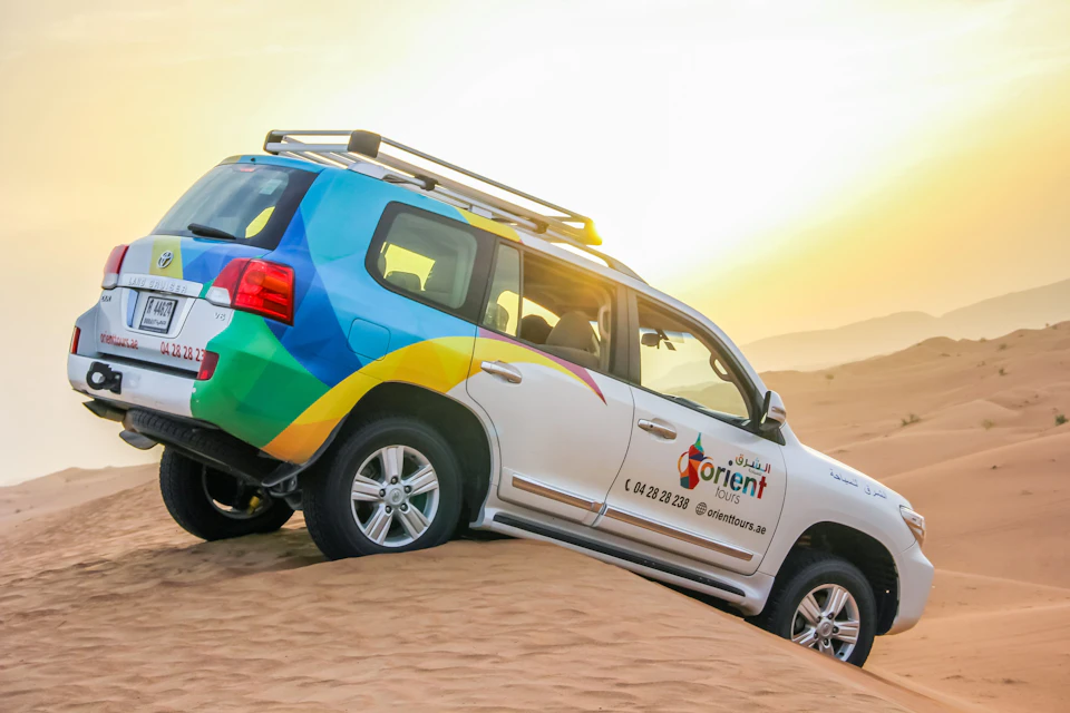 A colorful SUV with "Orient Tours" branding drives up a sand dune in a desert at sunset.