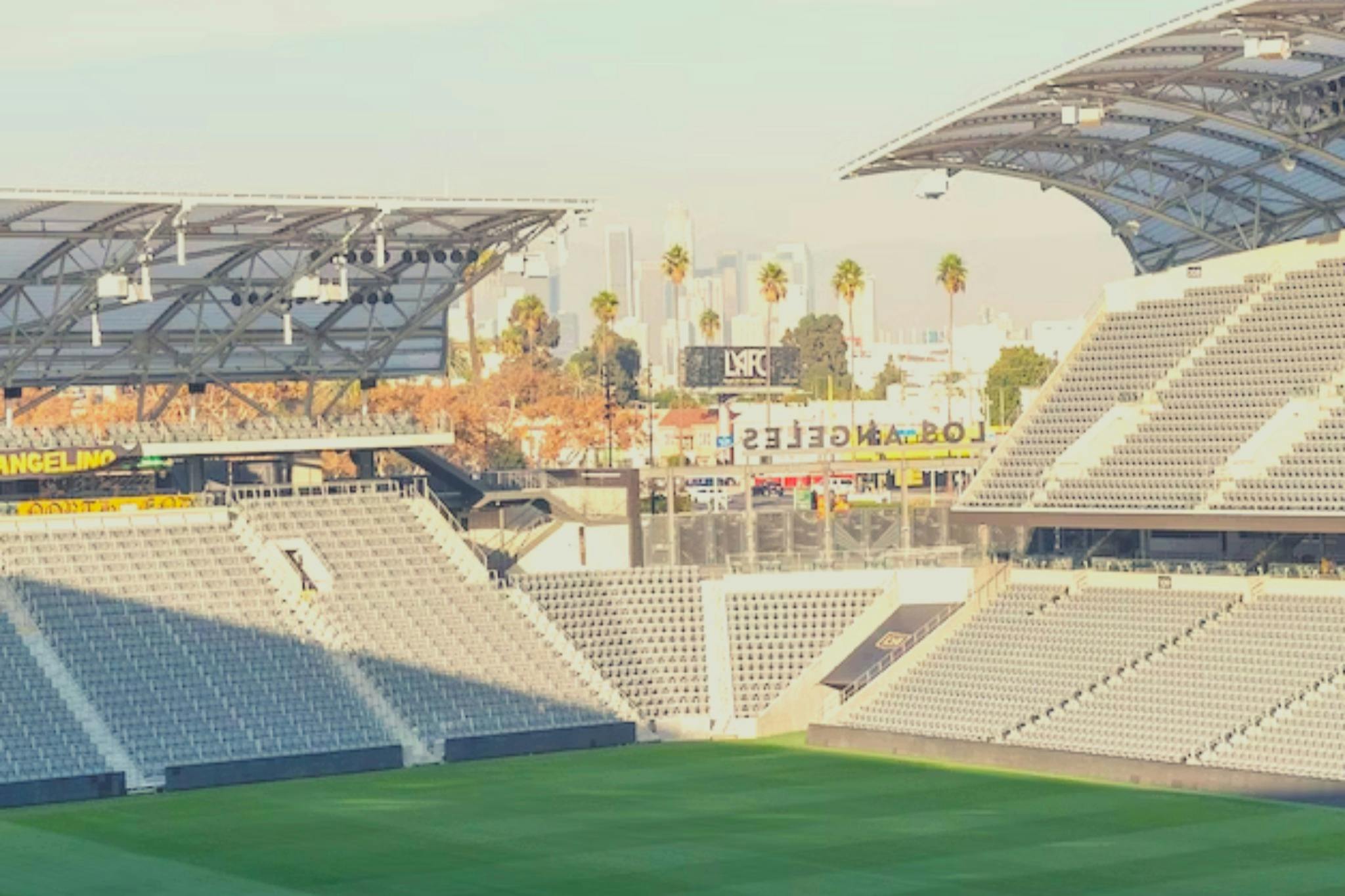 Empty stadium with gray seating, large covered areas, green field, and a view of palm trees and city buildings in the background.