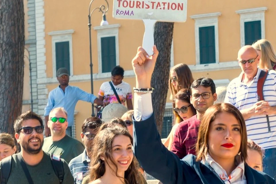A tour guide holding a sign leads a group of people through an outdoor urban area with historic architecture and trees.