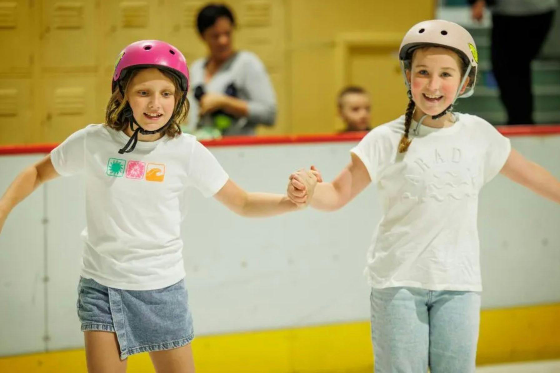 Two young girls wearing helmets hold hands and smile while roller skating indoors, with blurry people in the background.