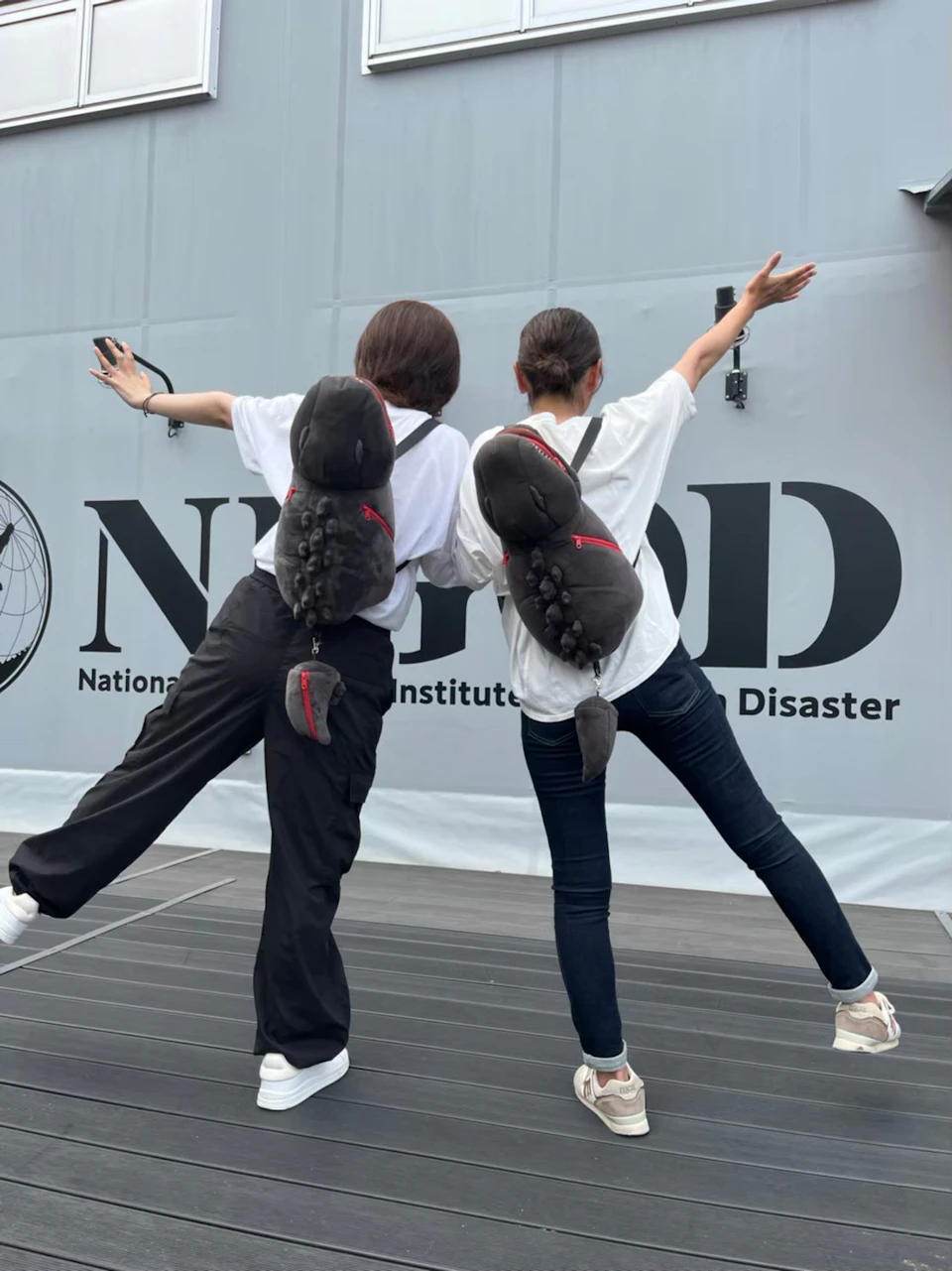 Two individuals wearing white shirts and plush animal-themed backpacks pose with arms outstretched in front of a modern building.