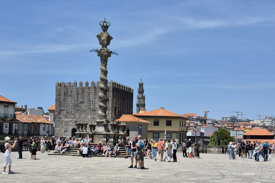 A historic square with people sitting and walking, a twisted stone column, and old buildings under a clear blue sky.