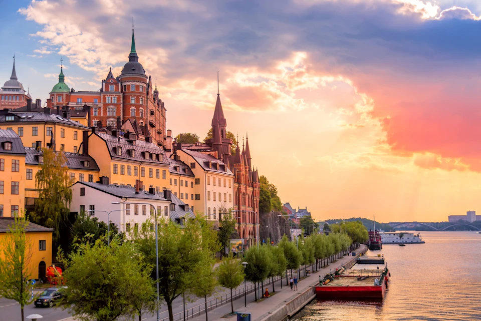 A scenic view of colorful buildings along a waterfront promenade with trees, under a vibrant sunset sky.
