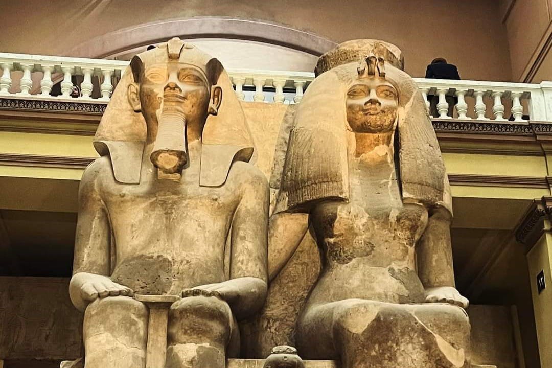 两座巨大的古埃及雕像，描绘了一位坐着的法老和一个戴着精致头饰的人物，在室内展出。