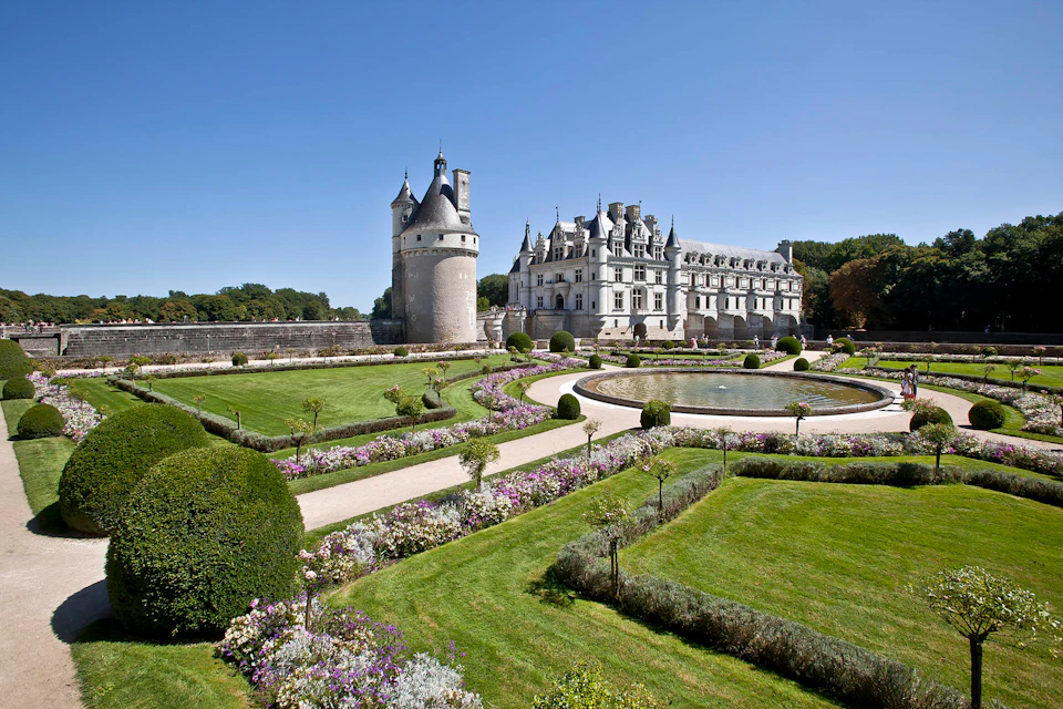 A historic castle with conical towers is surrounded by manicured gardens with curved pathways, symmetrically arranged greenery, and flowerbeds.
