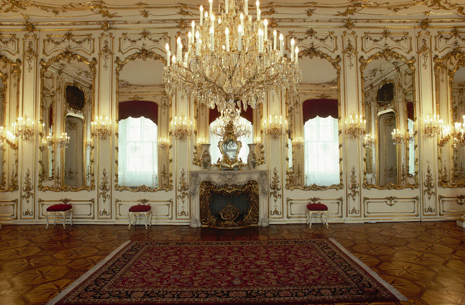 A lavish, ornate room with a large chandelier, intricate gold detailing, mirrors, a fireplace, and red upholstered stools.