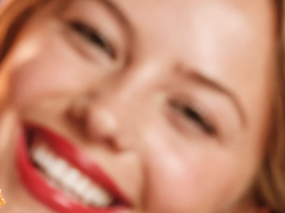 Smiling woman holding two large cheeseburgers with lettuce, bacon, and melted cheese. Neon sign in the background.