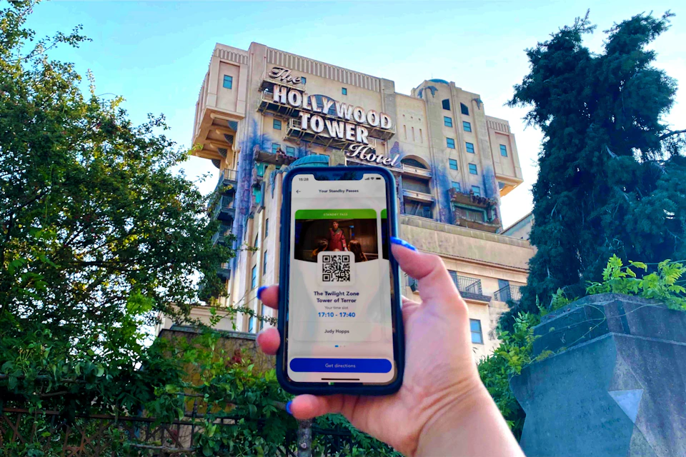 A hand holding a phone displaying a pass for The Twilight Zone Tower of Terror in front of the Hollywood Tower Hotel.