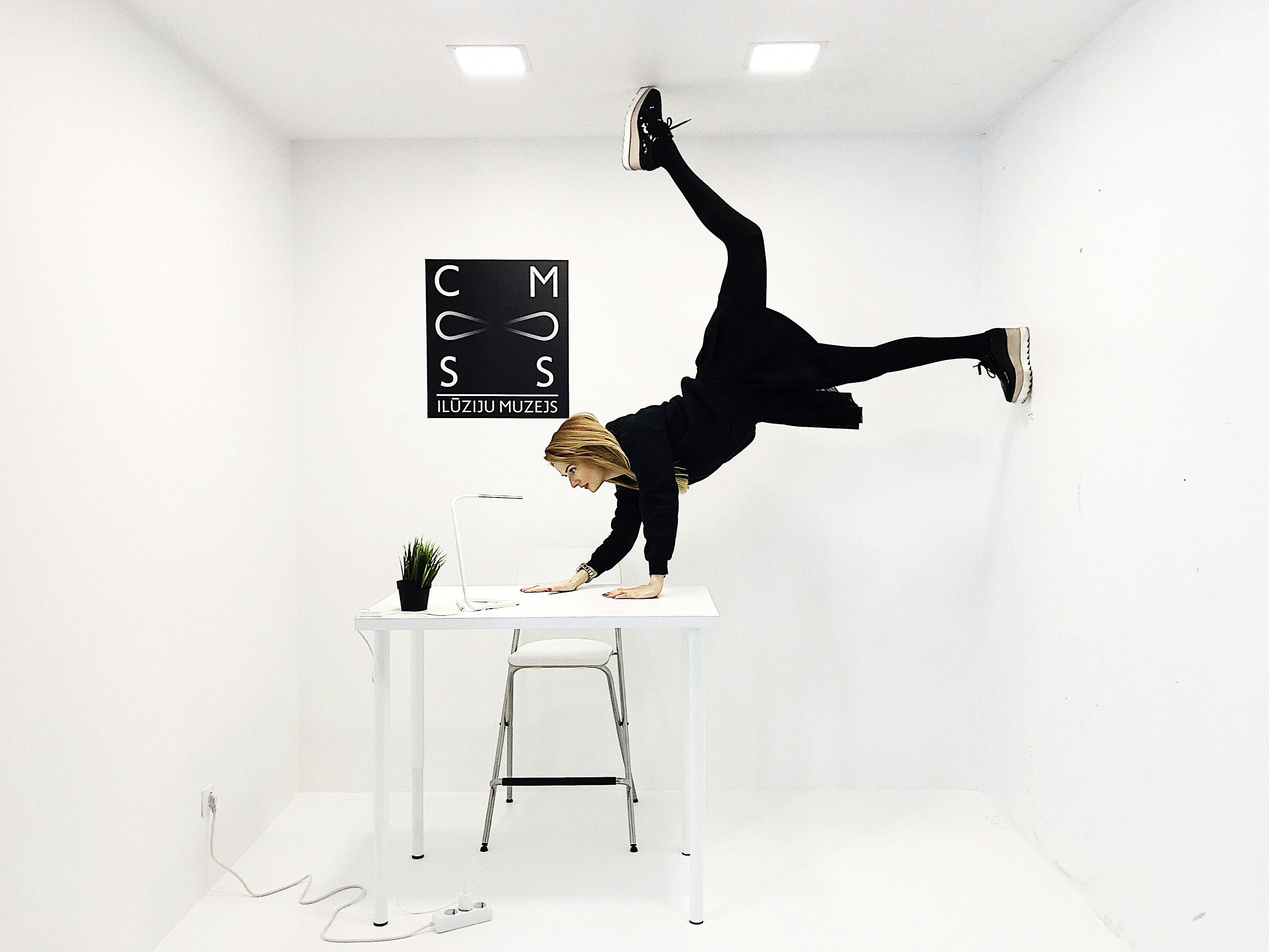 一个女人在全白房间的桌子上做倒立，墙上挂着一个单色的幻觉博物馆标志。