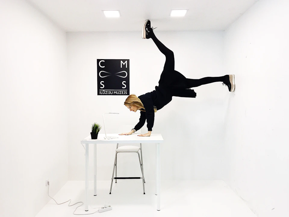 A woman does a handstand on a table in an all-white room, with a monochrome Illusions Museum sign on the wall.
